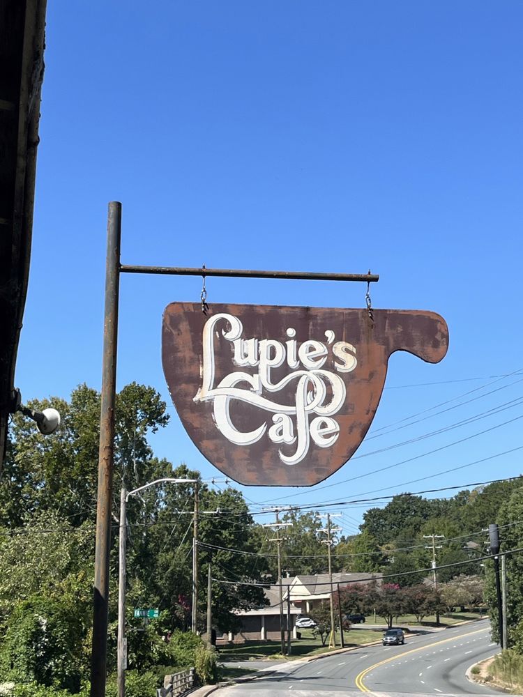The hanging sign on Monroe Road: a rusted steel coffee cup with Lupie's Cafe hand lettered in white.