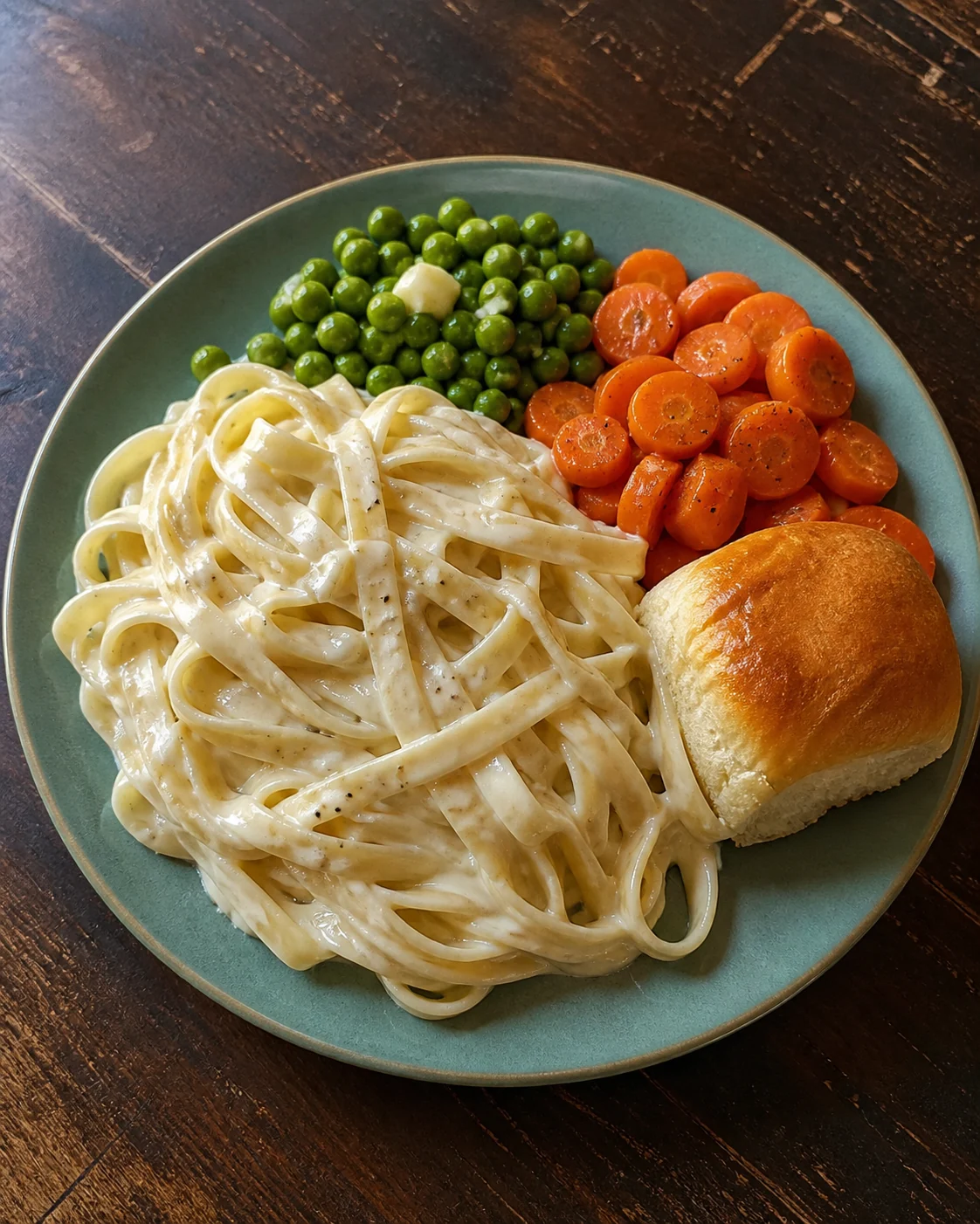 Fettuccine alfredo on a teal plate with buttered peas, carrots and a dinner roll.