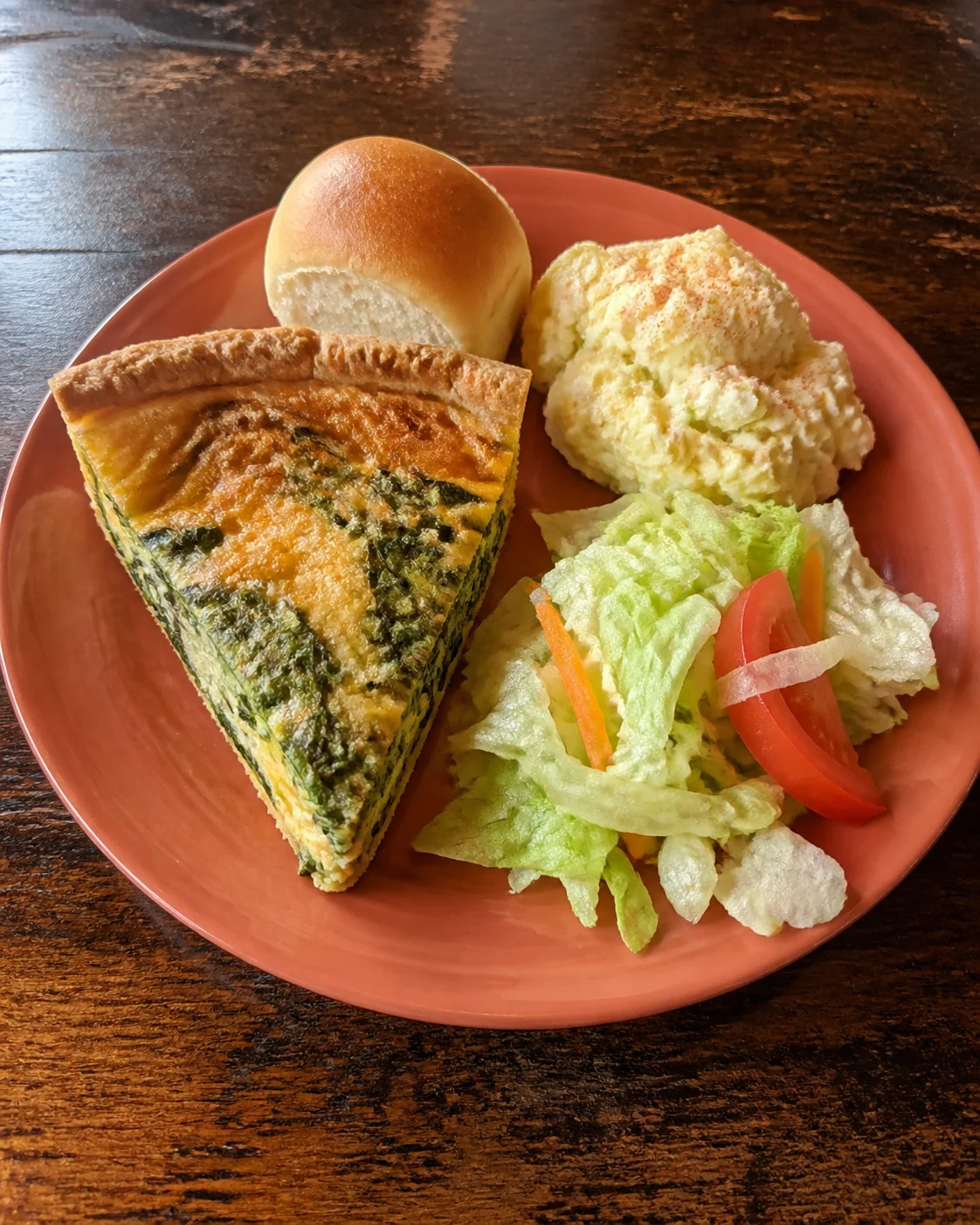 A wedge of spinach quiche on a terracotta plate with potato salad, a roll and a green salad.